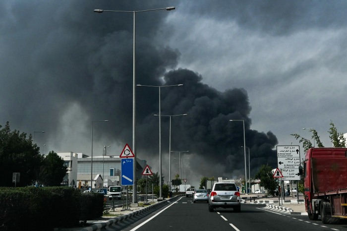 Vehicles driving on a street with a large plume of dark smoke rising in the background.